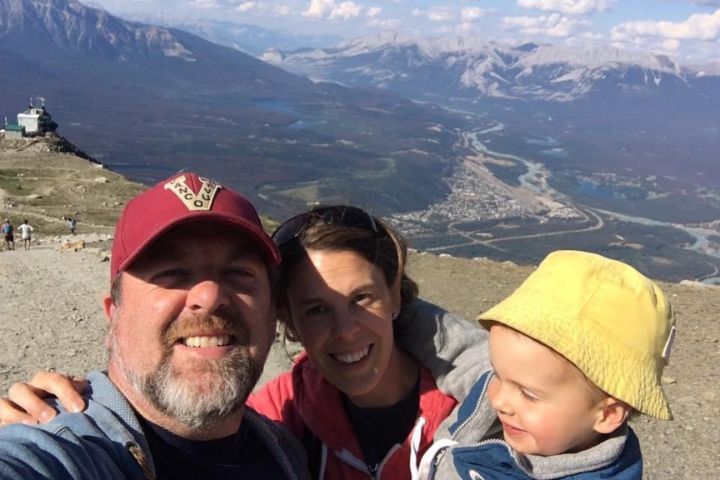 A family with a small baby wearing a yellow hat in front of a mountainous landscape.