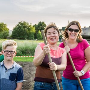In For Care: Local cooperation gardens - in 4 neighbourhoods, residents would like to have a common garden, a piece of land, where they can garden together and where they can meet each other. Two women and a boy in a garden holding gardening tools, smiling at the camera.