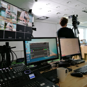 COM³: together despite distance. During the Covid-19 pandemic, COM³ helped local SMEs stay connected with their customers.With the support of COM³, companies showcased and promoted their products and services professionally using an online communication platform. Screens next to each other in the forefront; a man with a videocamera in the background.