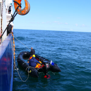 North Sea Wrecks: Where are the wrecks and ammunition in the North Sea? What dangers do they pose to people and the environment? What is the story behind them? We want to answer these questions. Sampling helps us to do this. Photo: Cornelia Riml A small rubber boat with people in it next to a larger ship, with deep blue sea and light blue sky.